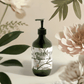 Ginger & Smoky Cardamom Hand-Body Wash bottle amidst flowers and leaves.